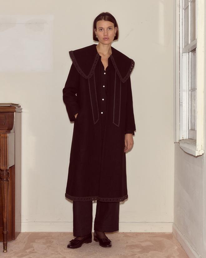 A woman stands by a window in a neutral room, wearing the Dover Coat—crafted from 100% wool with a wide collar and antique pearl buttons—paired with black pants and shoes.