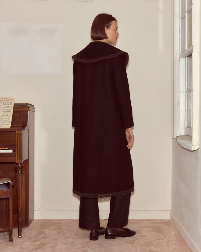 A person wearing the Dover Coat—a long, 100% wool formal coat with antique pearl buttons—stands indoors near a piano, facing away from the camera.