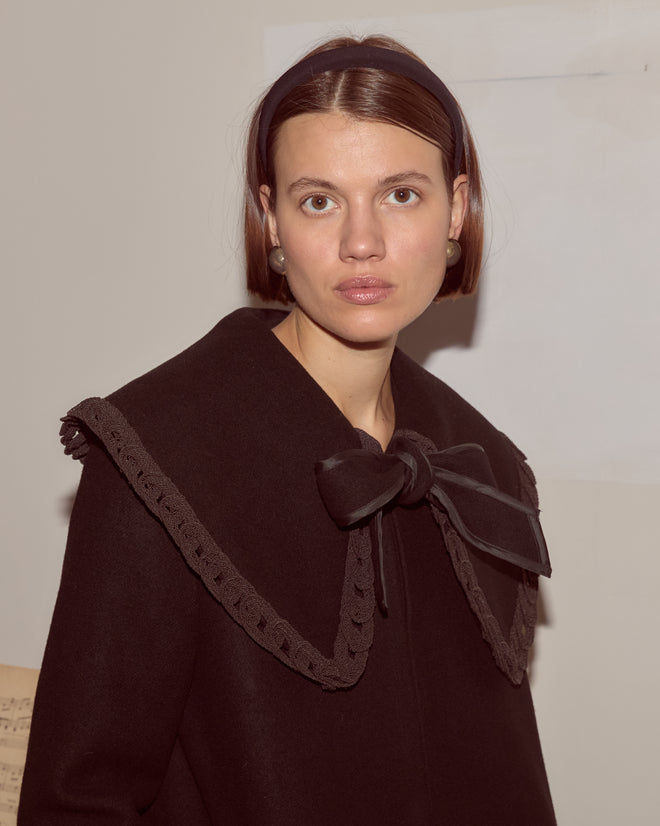 A woman in the Dover Coat, featuring antique pearl buttons and a large bow collar, paired with a headband, stands indoors against a plain background.
