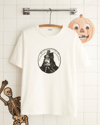 The Dracula Tee showcases an artistic depiction of a historical figure, beautifully set against a tiled wall decorated with pumpkins and skeletons, ideal for those looking for distinctive Halloween fashion.