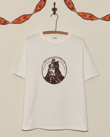 The Dracula Tee features a circular black illustration of a historical figure on a white shirt, hanging on a hook with festive garland above—ideal as a vintage or unique Halloween tee.