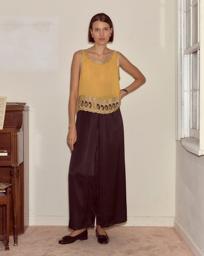 Woman in a yellow Ducat Tank and black wide-leg pants stands indoors by a window near a piano.