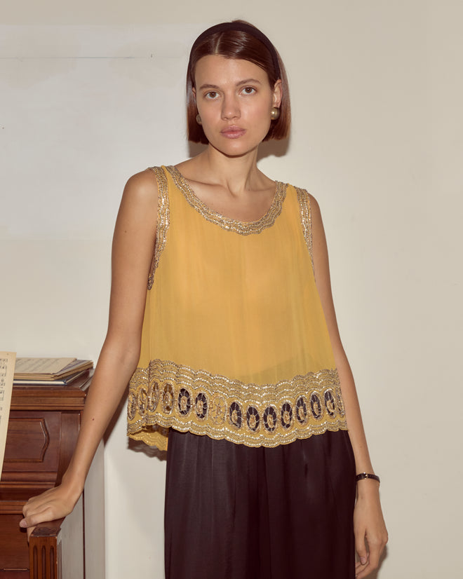 A woman in the Ducat Tank, a yellow silk top with a sequined hem, and black pants stands by a wooden piano, looking at the camera.