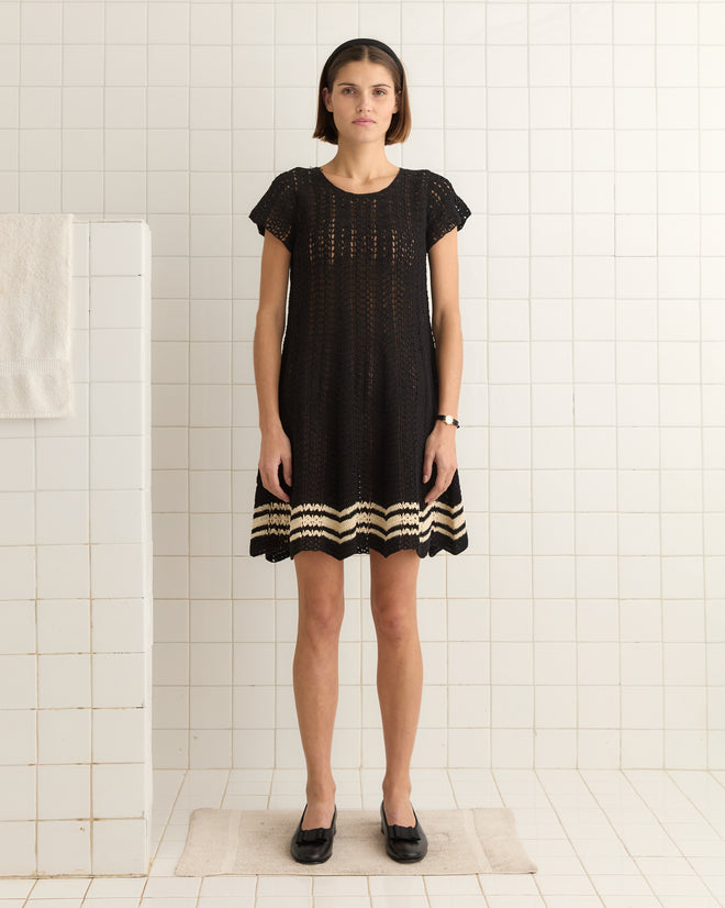 A person wearing the Duxbury Dress - Black stands on a bath mat in a tiled room, near a white towel hanging on a partition.