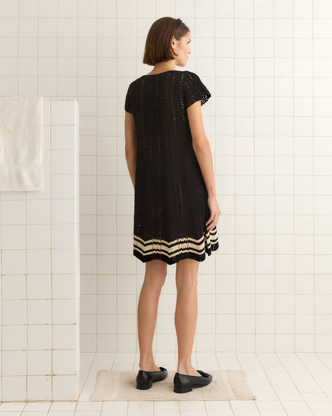 A woman in a Duxbury Dress - Black stands on a mat in the tiled bathroom, facing away.