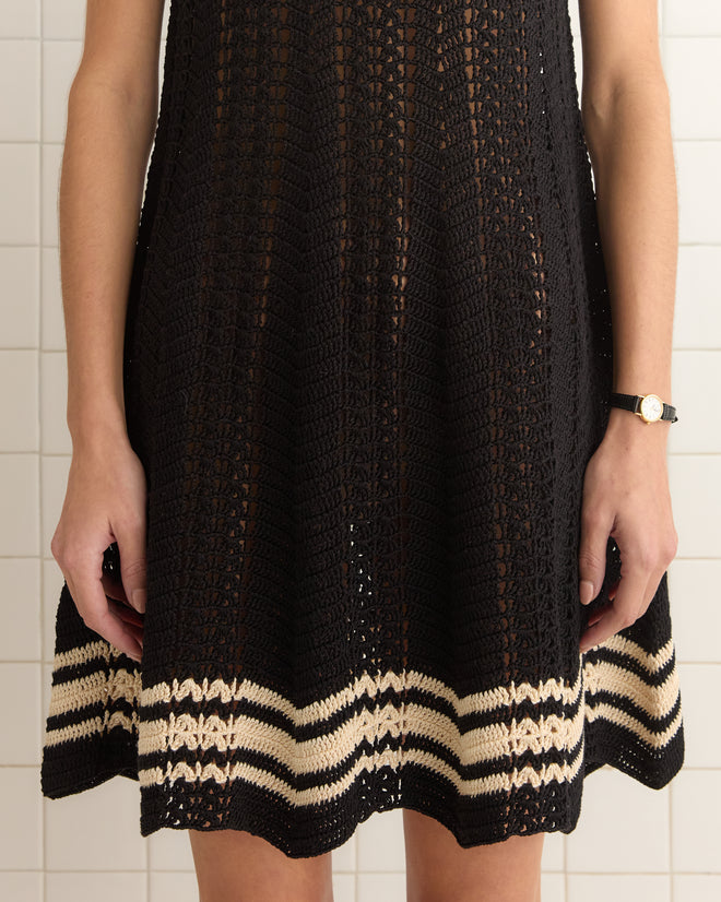 A person wearing the black Duxbury Dress, featuring a white-striped crochet pattern hem, stands against a tiled wall.