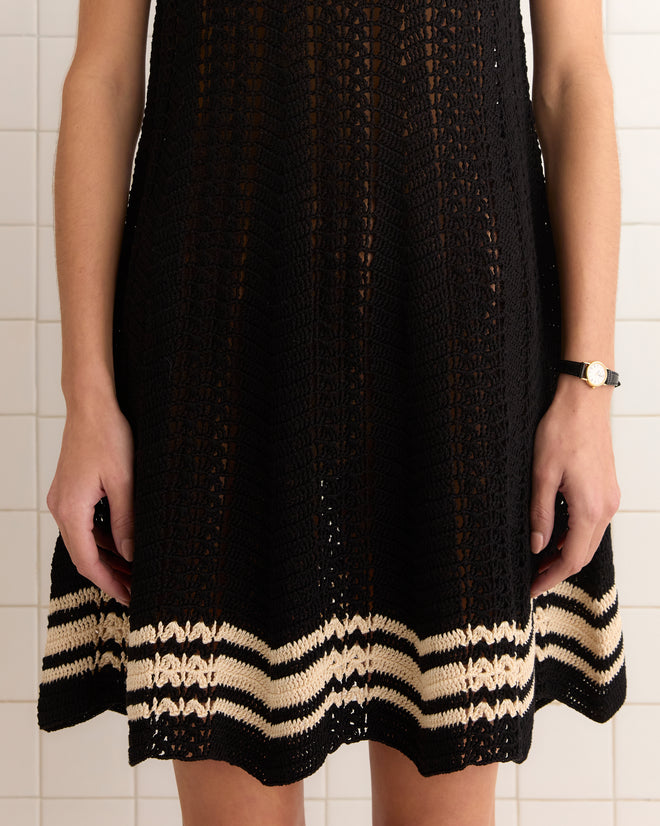 Wearing the Duxbury Dress in black, elegantly hand-crocheted with beige stripes at the hem, a person stands against a tiled wall.