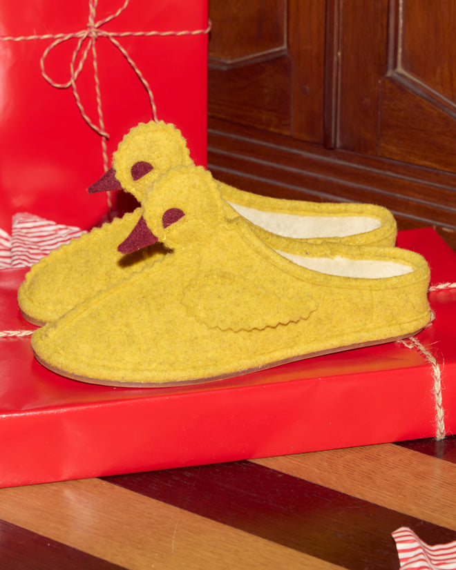 The Duck Slipper - Yellow Red, fleece-lined and chicken-shaped, rests atop red gift boxes on a wooden background.