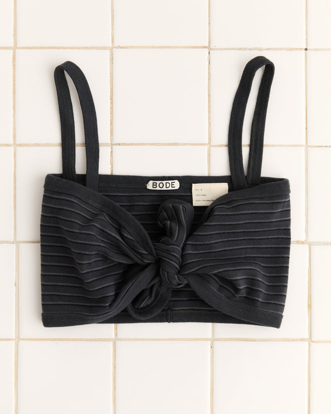The Ducky Bra, a black ribbed knit bralette featuring a tie-front detail and adjustable straps, is displayed against a white tiled background.