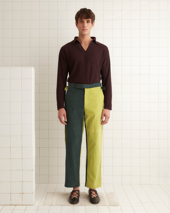 A pair of Duo Cord Trousers featuring a vintage-inspired design with one green leg and one yellow leg, hanging on a wooden hanger against a white wall.