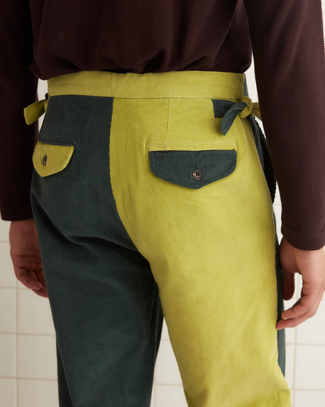 A pair of Duo Cord Trousers featuring a vintage-inspired design with one green leg and one yellow leg, hanging on a wooden hanger against a white wall.