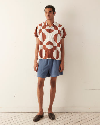 A person stands against a plain wall wearing the Duo Oval Patchwork Short Sleeve Shirt, featuring a brown and white geometric quilt top pattern, paired with blue shorts and brown slip-on shoes.