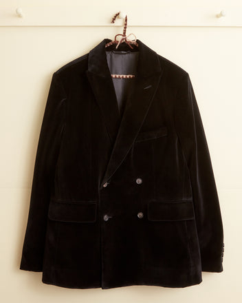 The Dusk Velvet Double-Breasted Suit Jacket for men, evoking the style of the 1950s-1960s, is hanging on a hook against a light-colored wall.