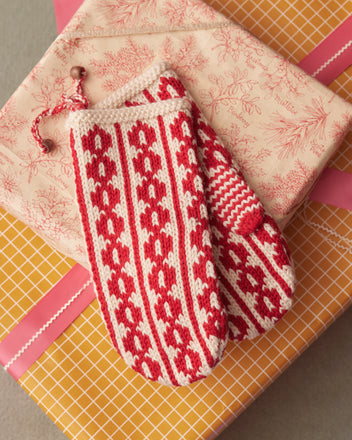 The Dutchess Mittens, crafted in a red and white knit from soft merino wool, rest on top of wrapped gifts adorned with pink ribbons and festive patterns.