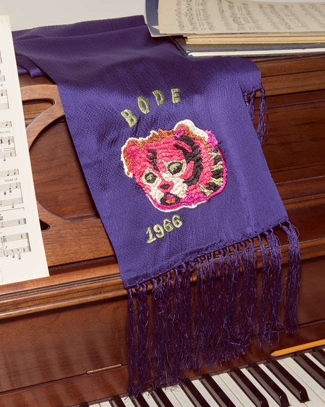 The Dream Souvenir Scarf—a purple, textured silk scarf with a tiger face and "BODE 1966" embroidery—drapes elegantly over a piano with sheet music, creating an Alice in Wonderland-inspired statement.