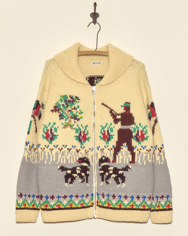 The Pheasant Cardigan, a cotton knitwear sweater, features colorful hunting and dog motifs on a beige and gray background, displayed hanging on a hook.