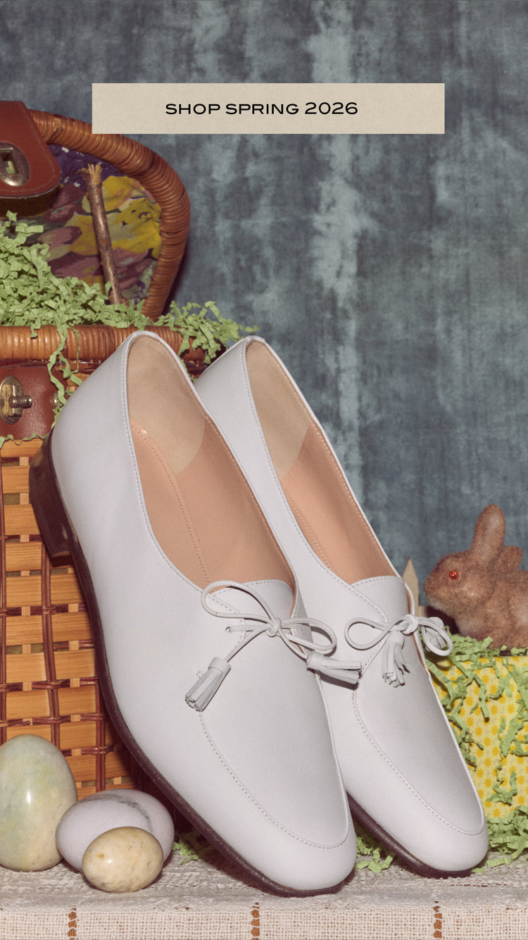 A pair of white tassel loafers sit on a lace covered table, surrounded by easter bunnies, eggs, and a wicker basket overflowing with green paper "grass".