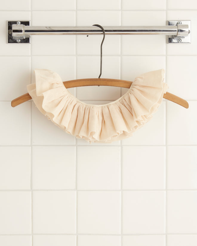 The Easy Ruff, a ruffled beige garment crafted from open-weave cotton, hangs elegantly on a wooden hanger suspended from a metal bar against a white tiled wall.