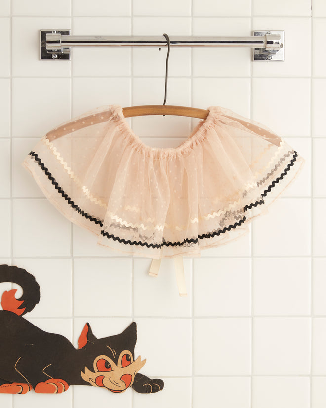 A sheer pink cape with black trim, similar to the Ecru Ribbon Collar from a 1950s costume, hangs on a wooden hanger. A cartoon cat illustration adorns the white tiled wall.