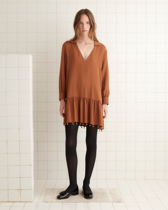 A woman wearing the Eddy Dress in a rust-brown color with a drop-waist silhouette, paired with black tights, stands in a tiled room with a white block next to her.