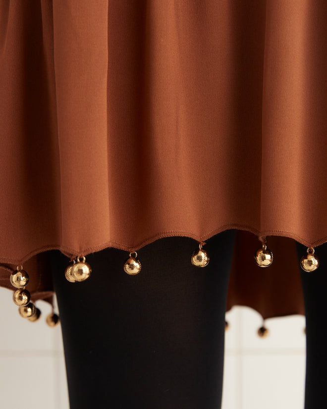 A woman wearing the Eddy Dress in a rust-brown color with a drop-waist silhouette, paired with black tights, stands in a tiled room with a white block next to her.