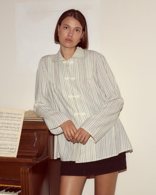 Woman wearing the Electric Light Blouse, a pale yellow stripe shirt, and a black skirt leans against a piano with sheet music, looking at the camera.