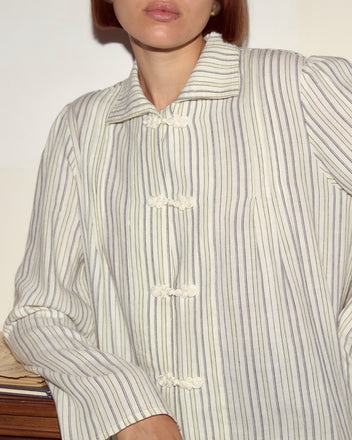 A woman in the Electric Light Blouse, a pale yellow striped button-up shirt, leans against a surface with her face partially out of frame.