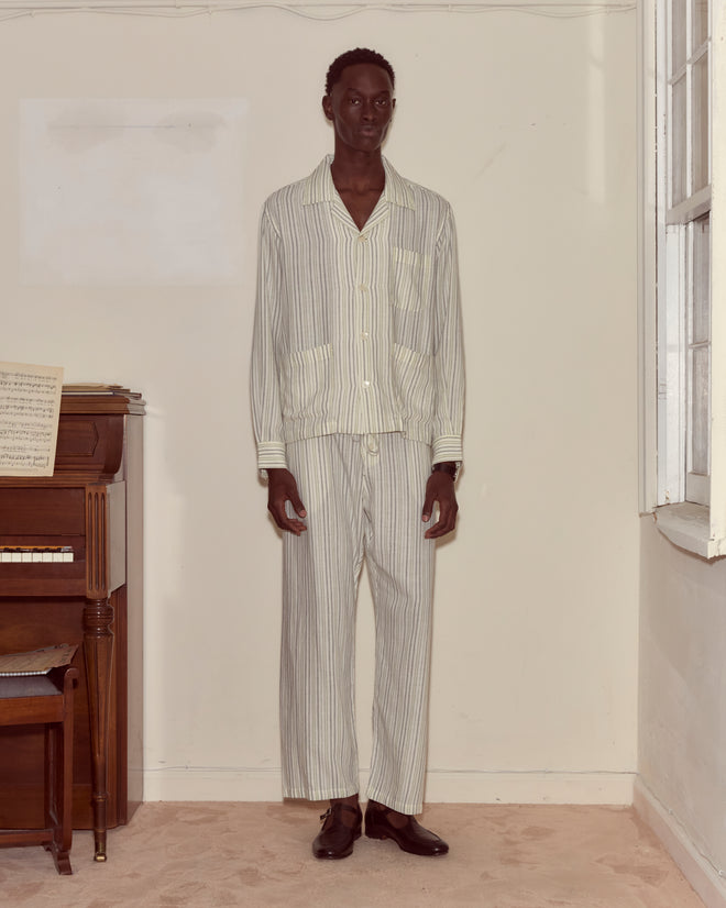 A man in Electric Light Long Sleeve pajamas stands indoors by a piano and window on a beige carpet.