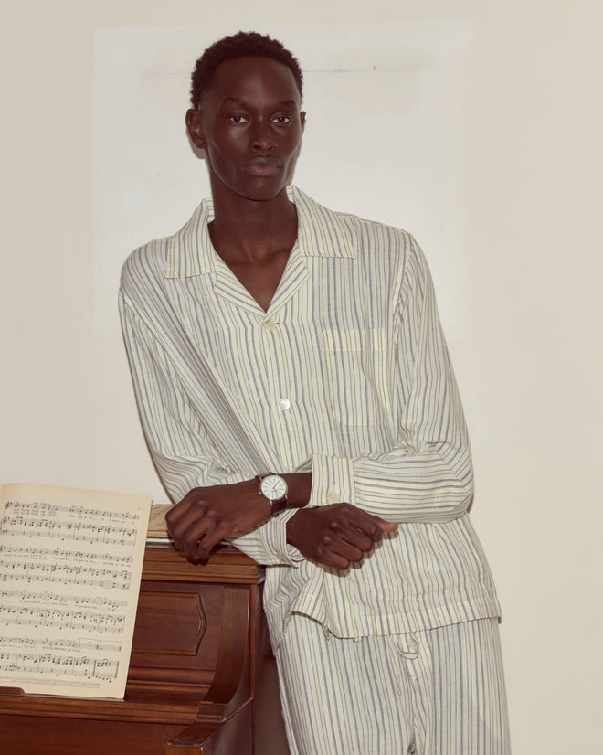 Wearing the Electric Light Long Sleeve, a person in yellow and black stripes leans on a piano with sheet music and a watch, looking to the side.