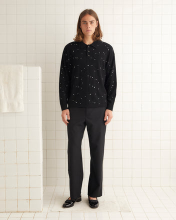 A person with long hair, wearing an Embellished Cycling Polo adorned with subtle rhinestones, black pants, and sandals, is standing in a tiled room.