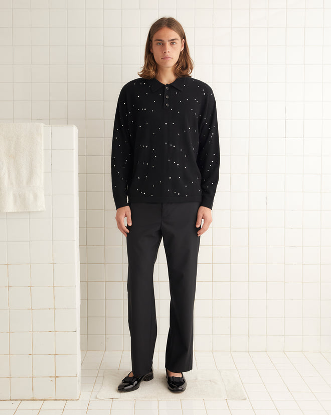 A person with long hair, wearing an Embellished Cycling Polo adorned with subtle rhinestones, black pants, and sandals, is standing in a tiled room.