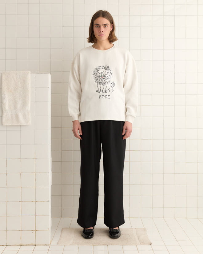 In a tiled room, the person stands confidently wearing the Embellished Lion Crewneck, a white sweater with a rhinestone lion motif and "BODE," paired with black pants and shoes.