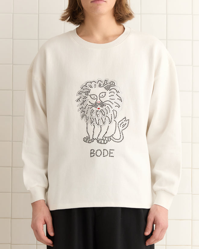 Against a tiled wall, a person wears the Embellished Lion Crewneck, showcasing casual elegance with its lion motif and "BODE" emblem.