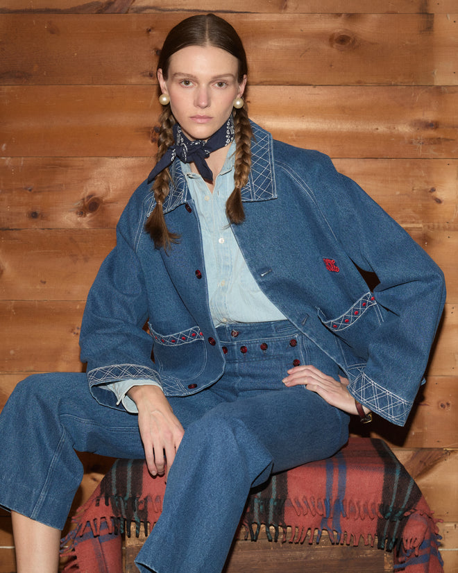 A woman with braided hair wears an Embroidered Denim Jacket as she sits on a blanket-covered bench against a wooden wall.