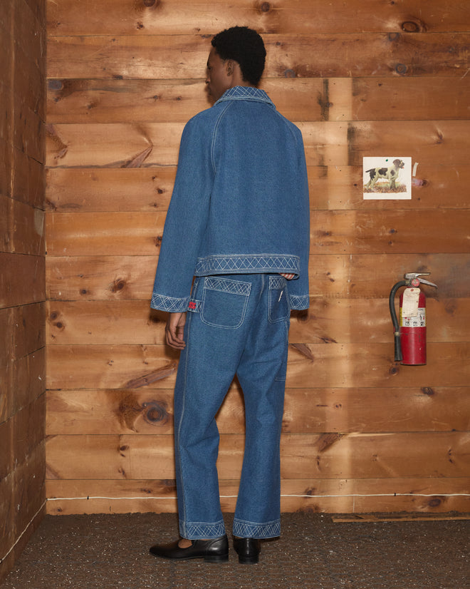 Wearing the Embroidered Denim Jacket, the person stands facing a wooden wall near a fire extinguisher and a small wall photo.