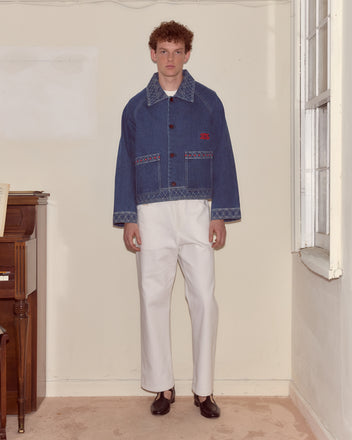 A man in an Embroidered Denim Jacket and white pants stands indoors by a window and a wooden piano.