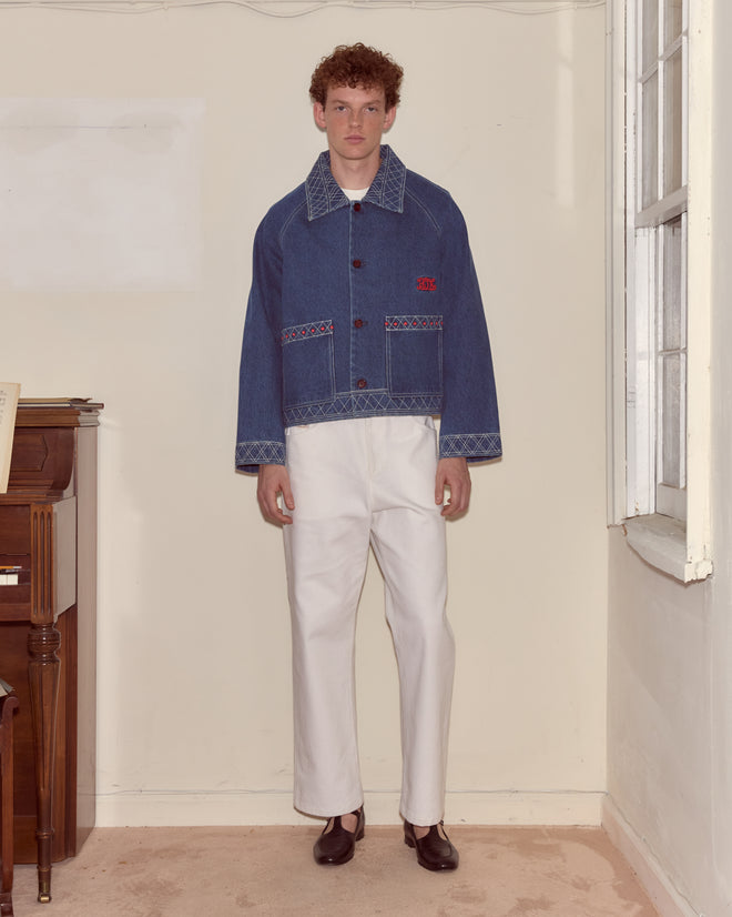 A man in an Embroidered Denim Jacket and white pants stands indoors by a window and a wooden piano.