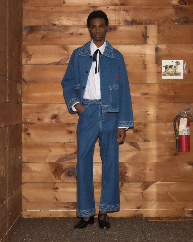A person wearing the Embroidered Denim Jacket, paired with matching denim pants featuring workwear details, a white shirt, and a bolo tie, stands next to a wood wall with a fire extinguisher.