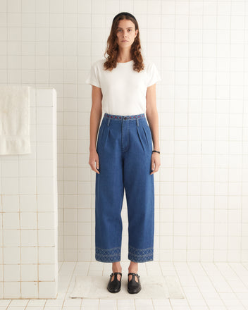 A person stands in a tiled room wearing a white T-shirt, Embroidered Denim Murray Trousers with an embroidered waistband, and black shoes.