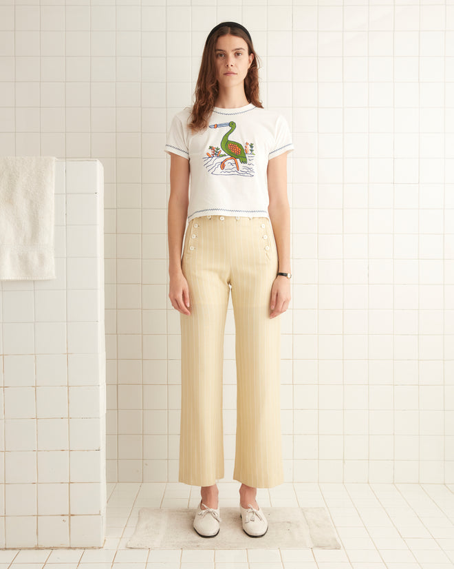 A woman stands in a tiled bathroom wearing the Embroidered Heron Tee, a white t-shirt made from 100% cotton featuring a colorful graphic, paired with beige striped pants. She is facing the camera with a neutral expression.