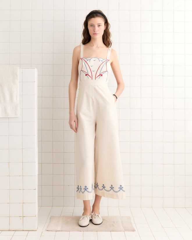 A woman in the Embroidered Sailboat Jumpsuit - White/Multi, featuring red and blue embroidery, stands in a tiled room with hands in pockets and wearing white shoes.