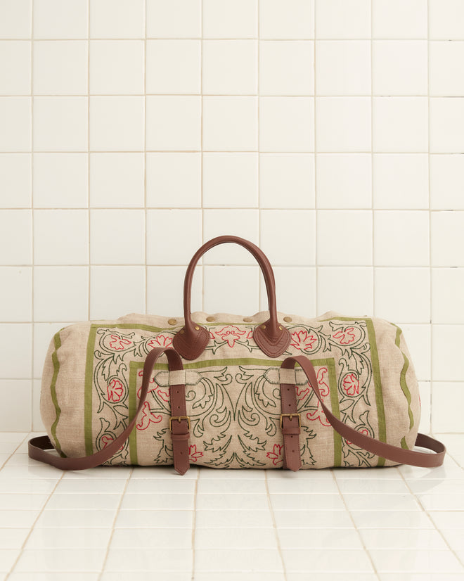 The Embroidered Trumpetflower Duffle Bag, featuring a beige and green pattern with brown leather straps, rests elegantly against a white tiled wall.