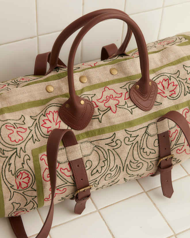 The Embroidered Trumpetflower Duffle Bag, featuring a beige and green pattern with brown leather straps, rests elegantly against a white tiled wall.