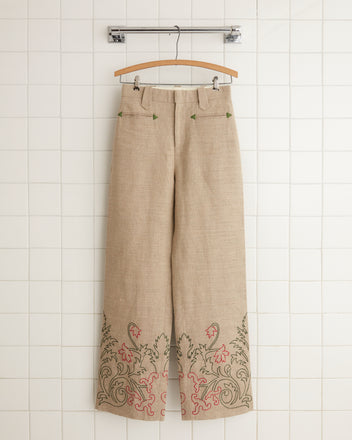 Embroidered Trumpetflower Murphy Trousers in beige, featuring wide-leg design and green and red floral embroidery on the hems, hanging on a hanger against a tiled wall.