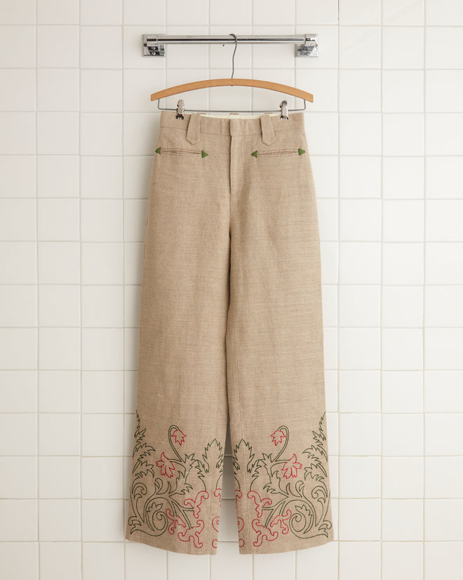 Embroidered Trumpetflower Murphy Trousers in beige, featuring wide-leg design and green and red floral embroidery on the hems, hanging on a hanger against a tiled wall.