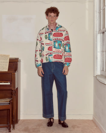 A man wearing the Enrichment Jacket and wide-leg jeans stands indoors beside a piano and a window.