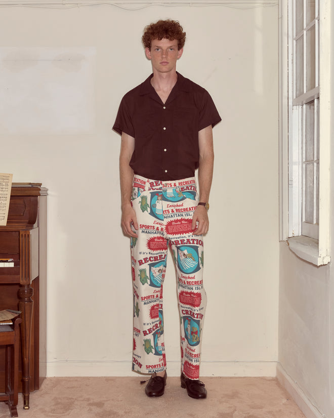 A young man in a brown shirt and Enrichment Knolly Brook Trousers stands indoors by a piano and window, looking at the camera.