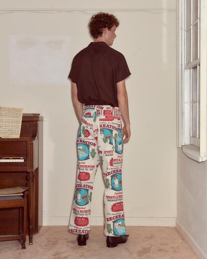 A person stands indoors facing away, wearing Enrichment Knolly Brook Trousers and a brown shirt near a piano and window.