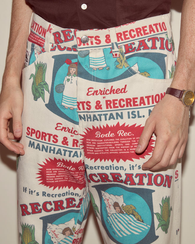 Wearing Enrichment Knolly Brook Trousers made from 100% cotton feedsack, featuring patterned vintage-inspired sports and recreation graphics, the person stands with hands by their sides.