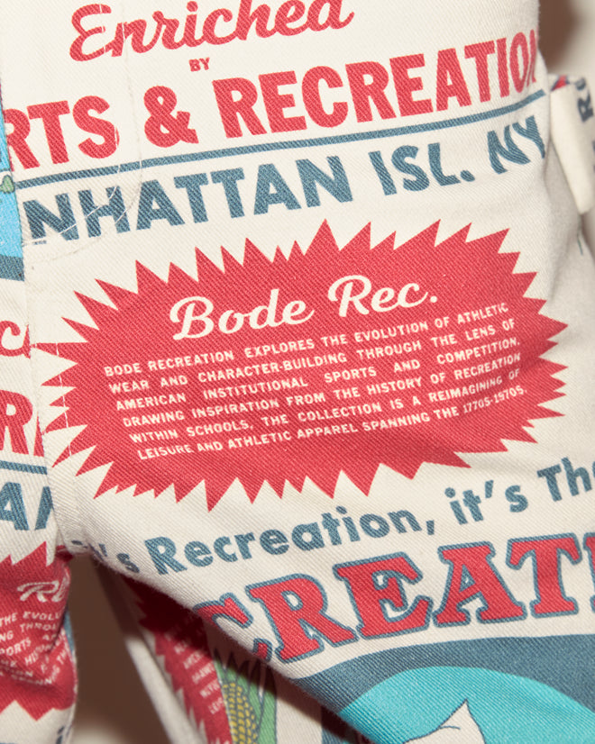 Close-up of fabric featuring red, blue, and white text inspired by athletic wear and recreation history. The 100% cotton Enrichment Knolly Brook Trousers recall classic feedsack workwear with a vintage feel.
